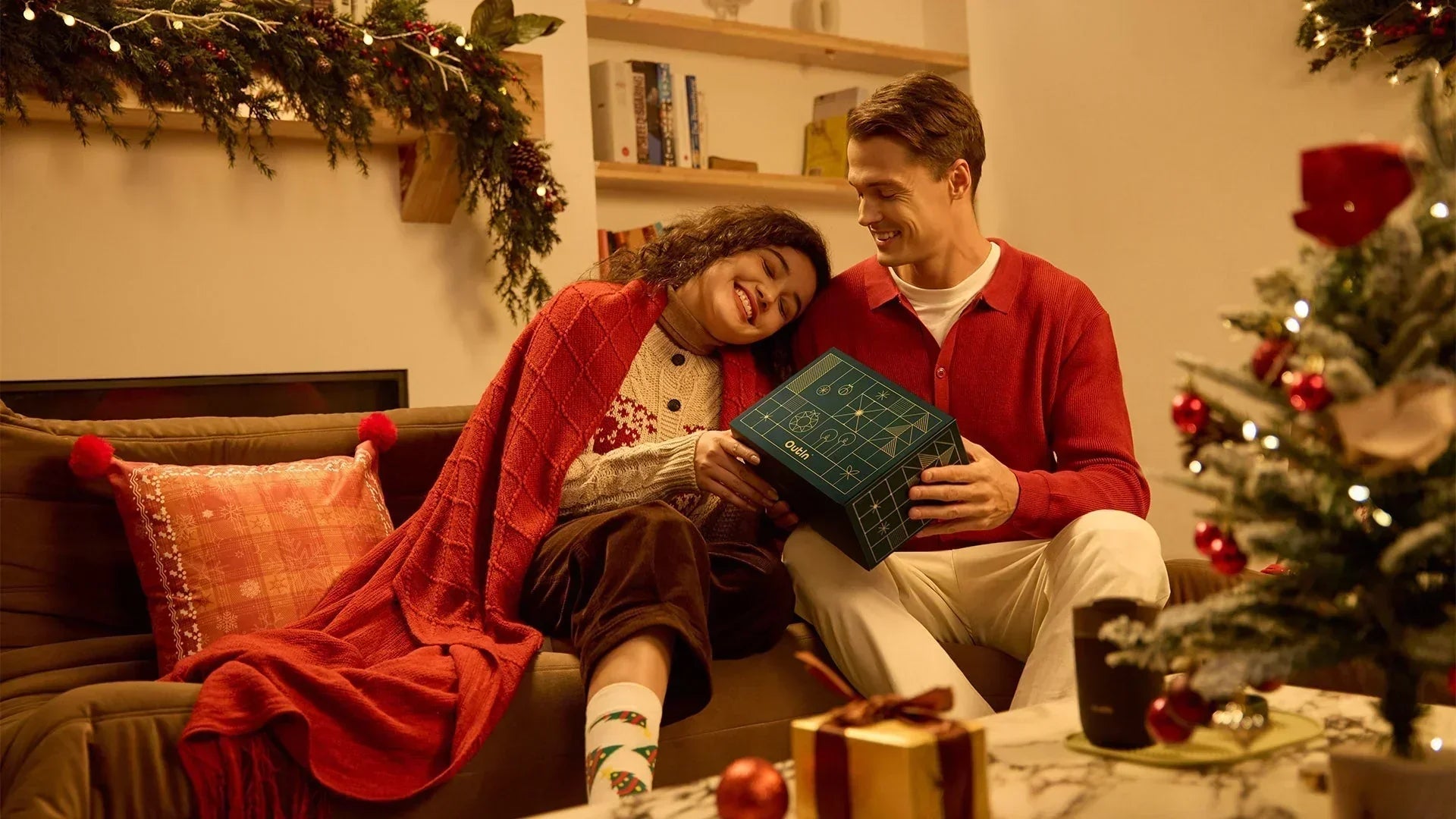 A cozy couple is sitting on a couch in a Christmas-decorated room, happily opening a dark green gift box labeled "Outin"