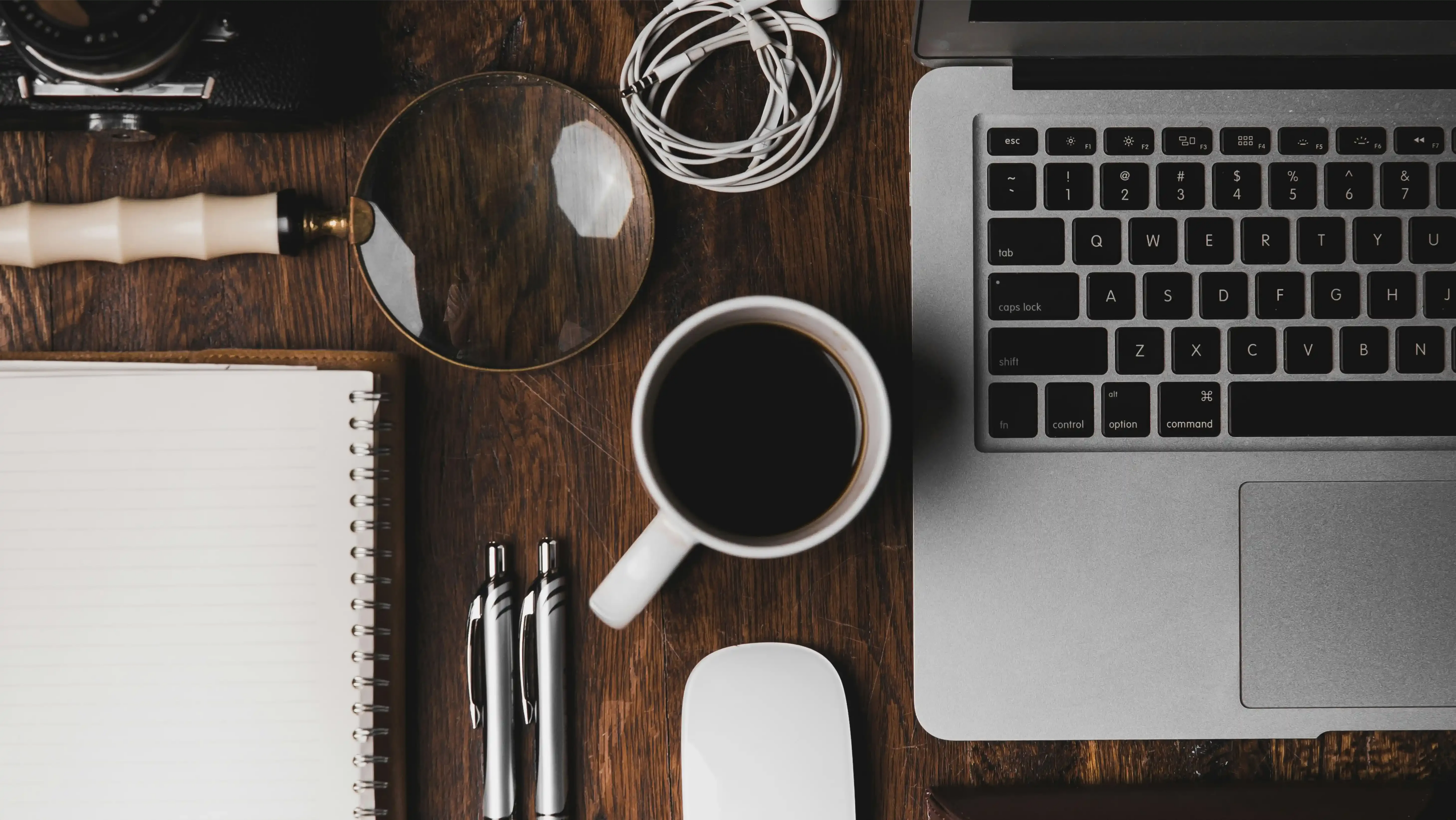 Minimalist office desk setup with laptop, coffee cup, white spiral notebook, magnifying glass, white earbuds and pens on wooden surface, captured from top view in moody lighting