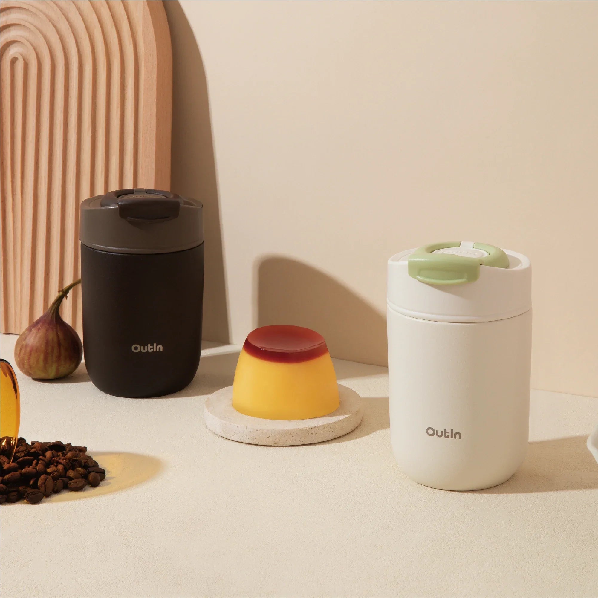 Two OutIn travel tumblers: matte dark brown with brown lid and matte cream with green lid. Pudding and coffee beans nearby.