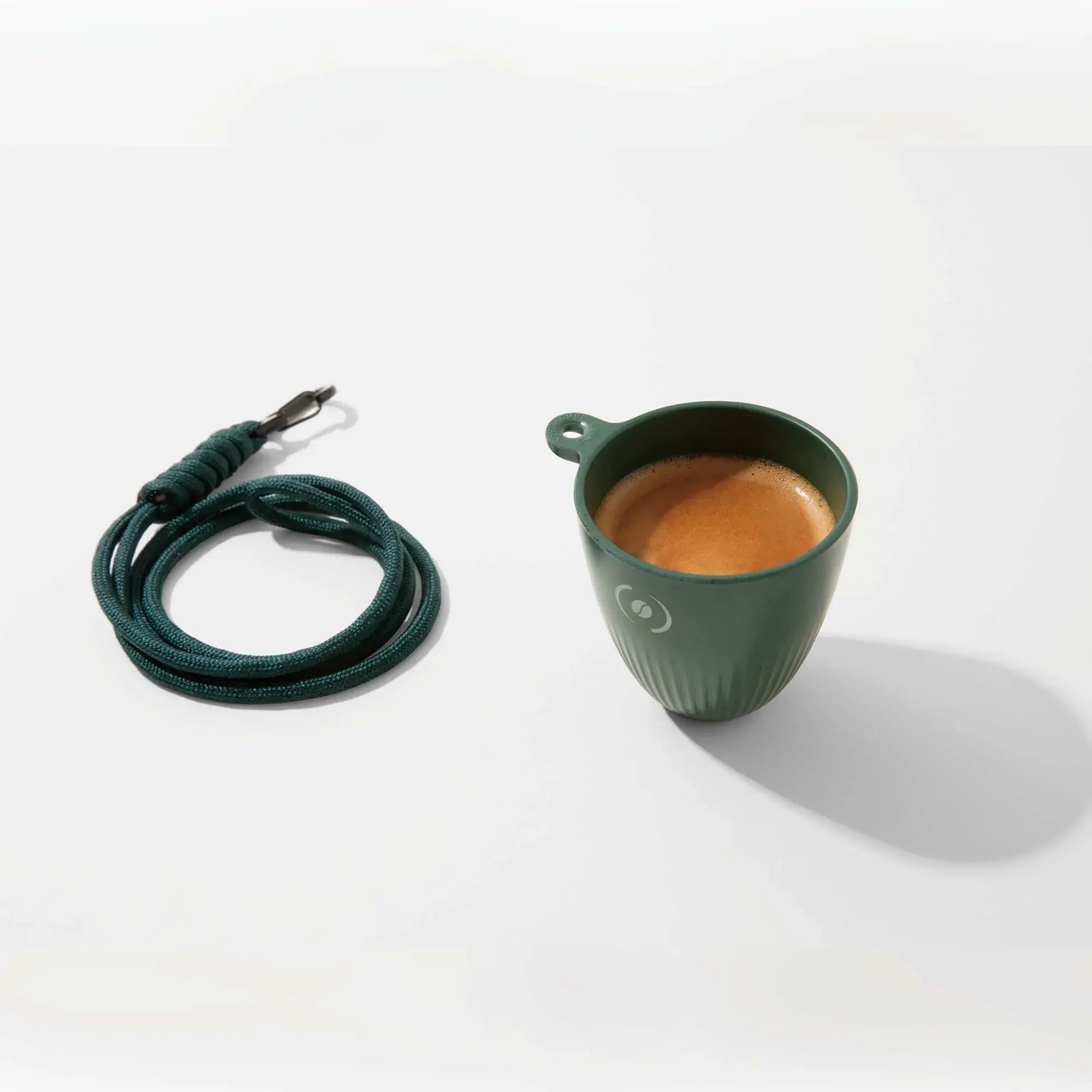 Dark green OutIn Rebrew ribbed espresso cup filled with coffee, with coiled lanyard and coffee bean logo.