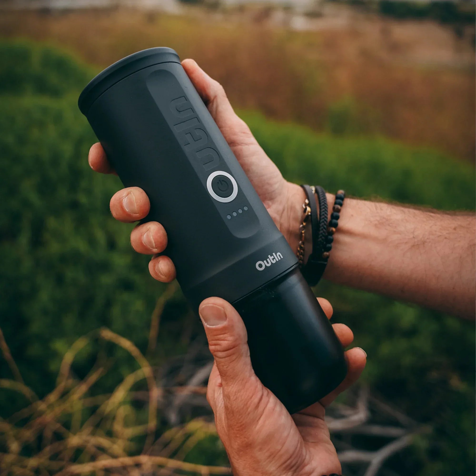 OutIn Nano Portable Espresso Machine in Space Grey held by hands outdoors, showing power button and LED indicators.