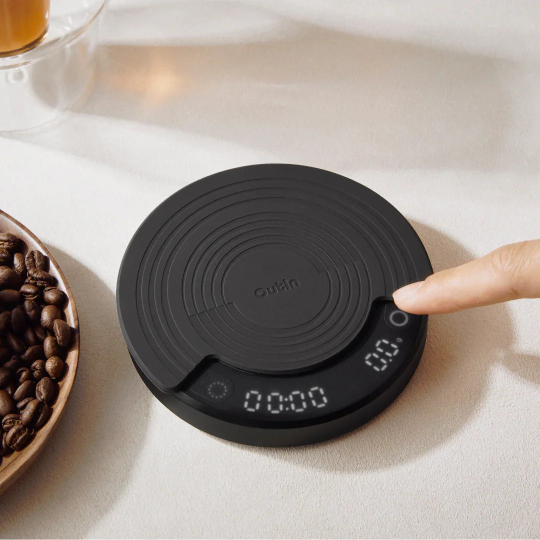 OutIn Claro coffee scale, dark gray, with digital display and side button activated. Features concentric circle texture.