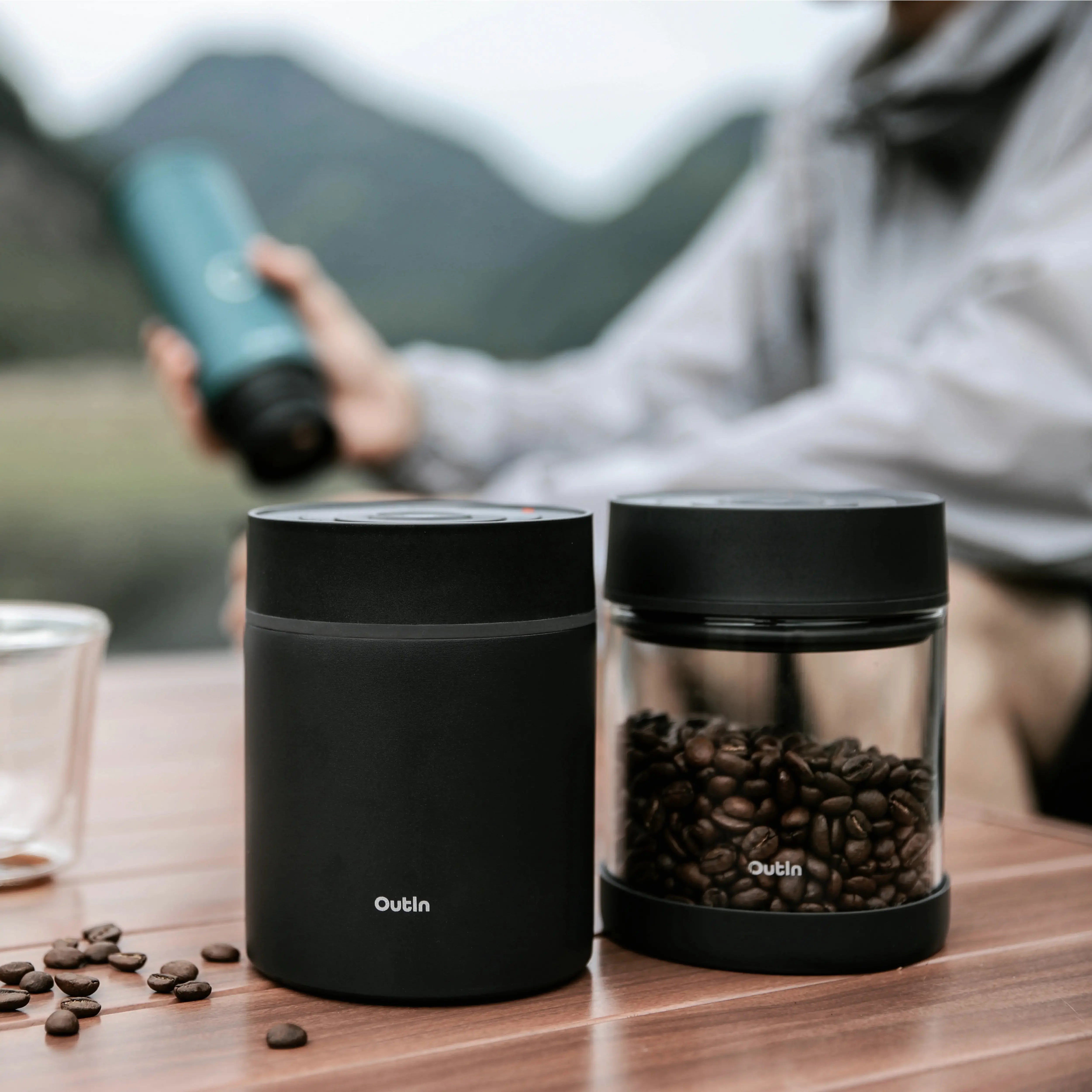 Two OutIn portable electric vacuum coffee canisters, black steel and glass with beans, on wooden table outdoors.
