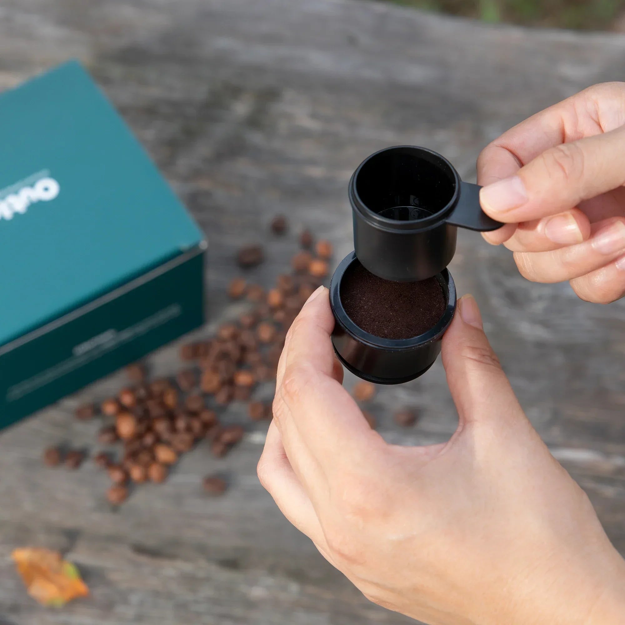 Hands using OutIn Nano ground coffee spoon to fill filter basket for portable espresso machine. Packaging and beans visible.