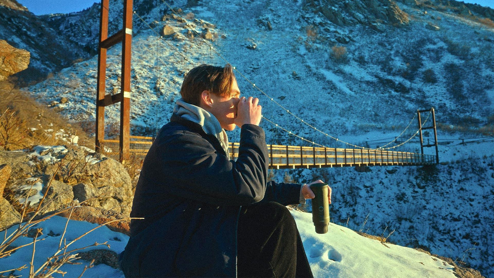 Person drinking coffee outdoors near a suspension bridge in a snowy mountainous area, holding an OutIn portable coffee maker