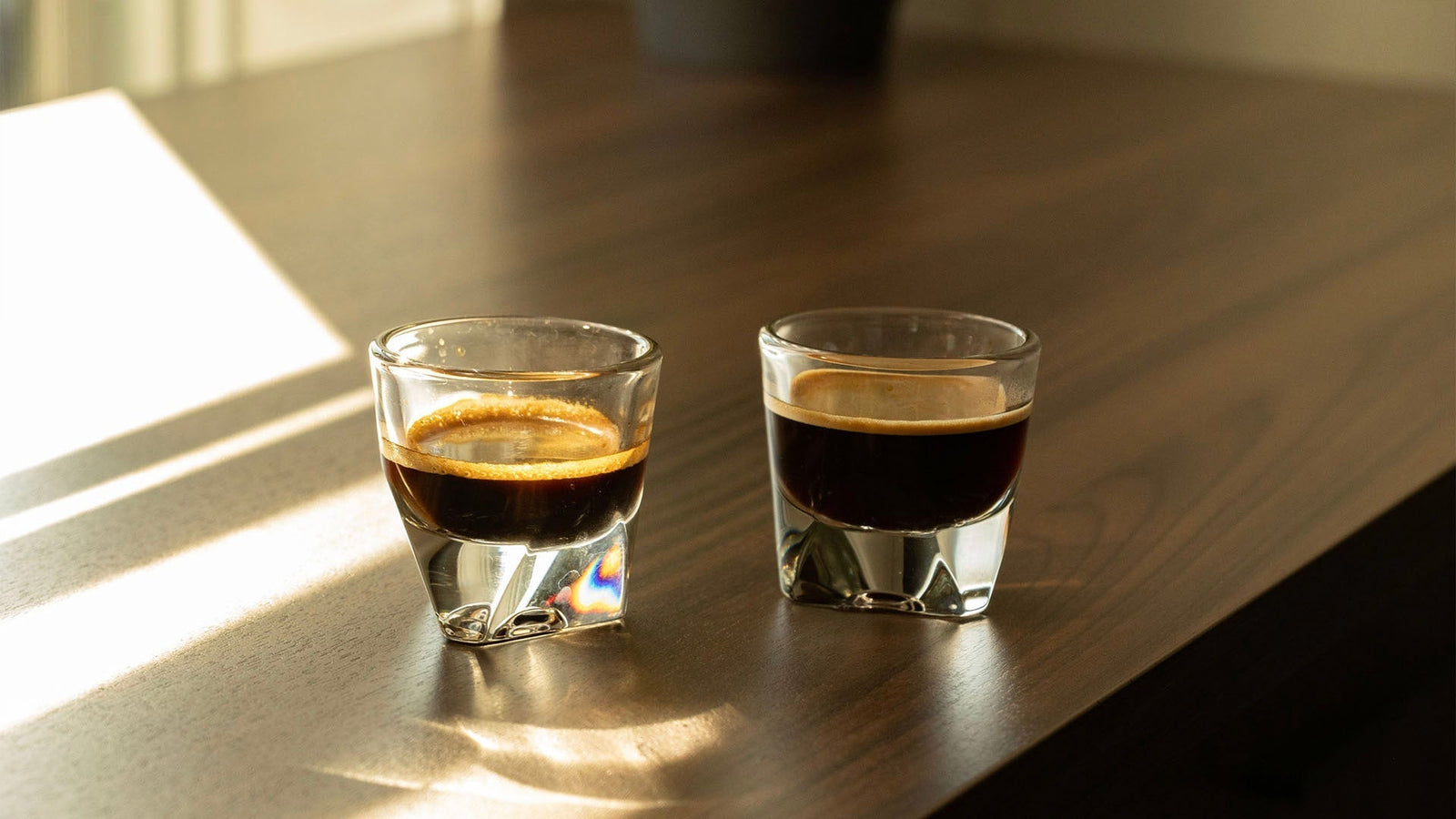 Two espresso shots in glass cups on a wooden table, bathed in warm sunlight with striped shadows