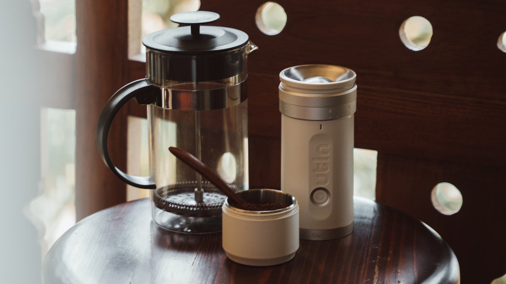 French press, Outin portable coffee grinder, and a container of freshly ground coffee on a wooden table, ready for brewing