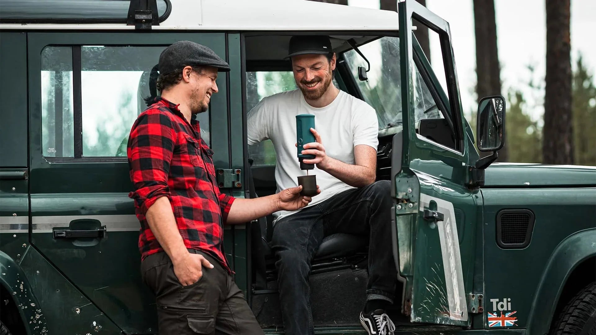 Two people enjoying coffee outdoors next to a green off-road vehicle with open door