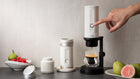 A finger pressing the power button on the white Outin espresso maker, with a shot of espresso dripping into a glass cup