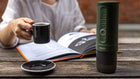 Outdoor scene with a hand holding a cup of espresso; the green Outin portable espresso maker rests on a book and wooden table