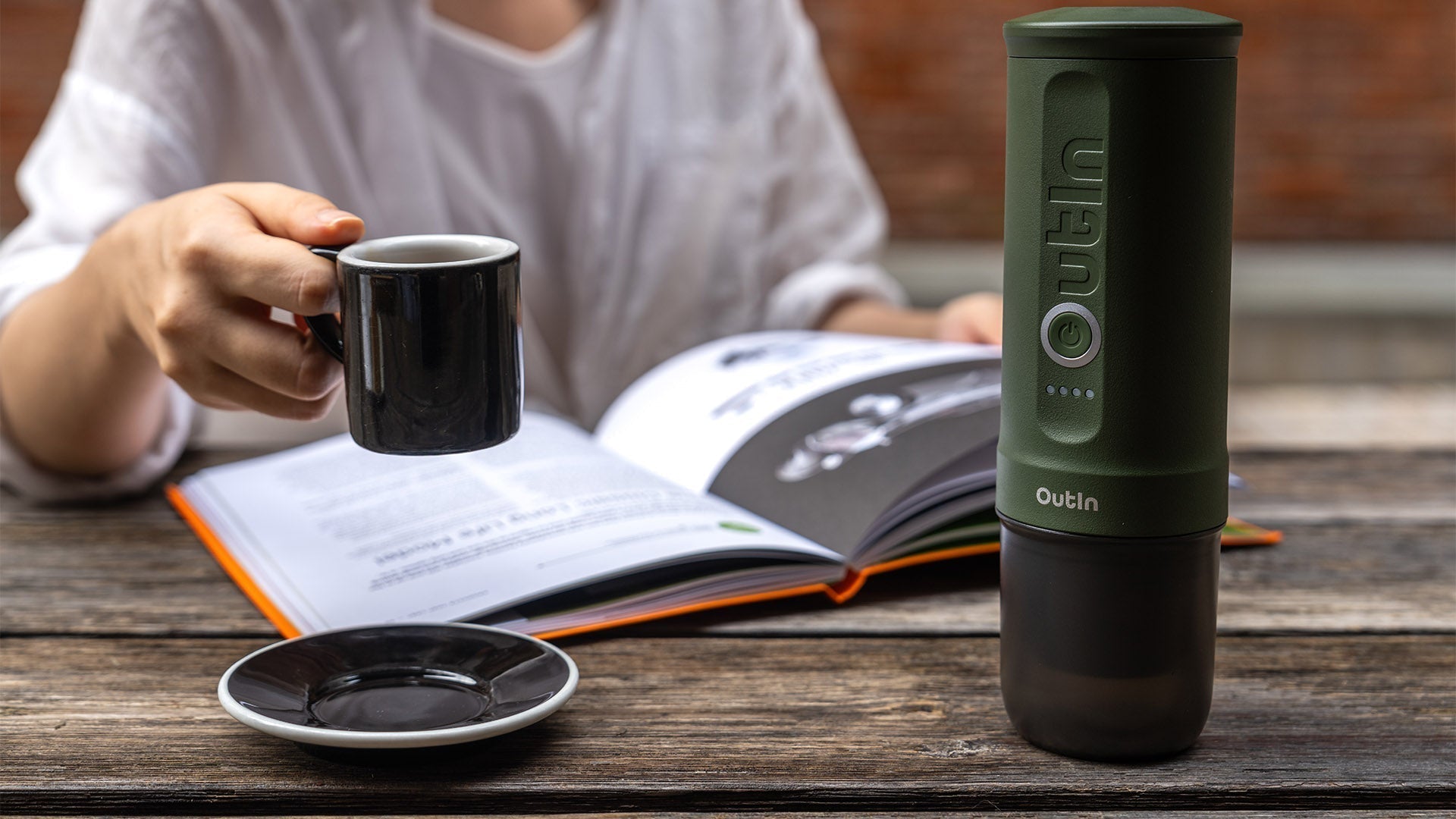 Outdoor scene with a hand holding a cup of espresso; the green Outin portable espresso maker rests on a book and wooden table