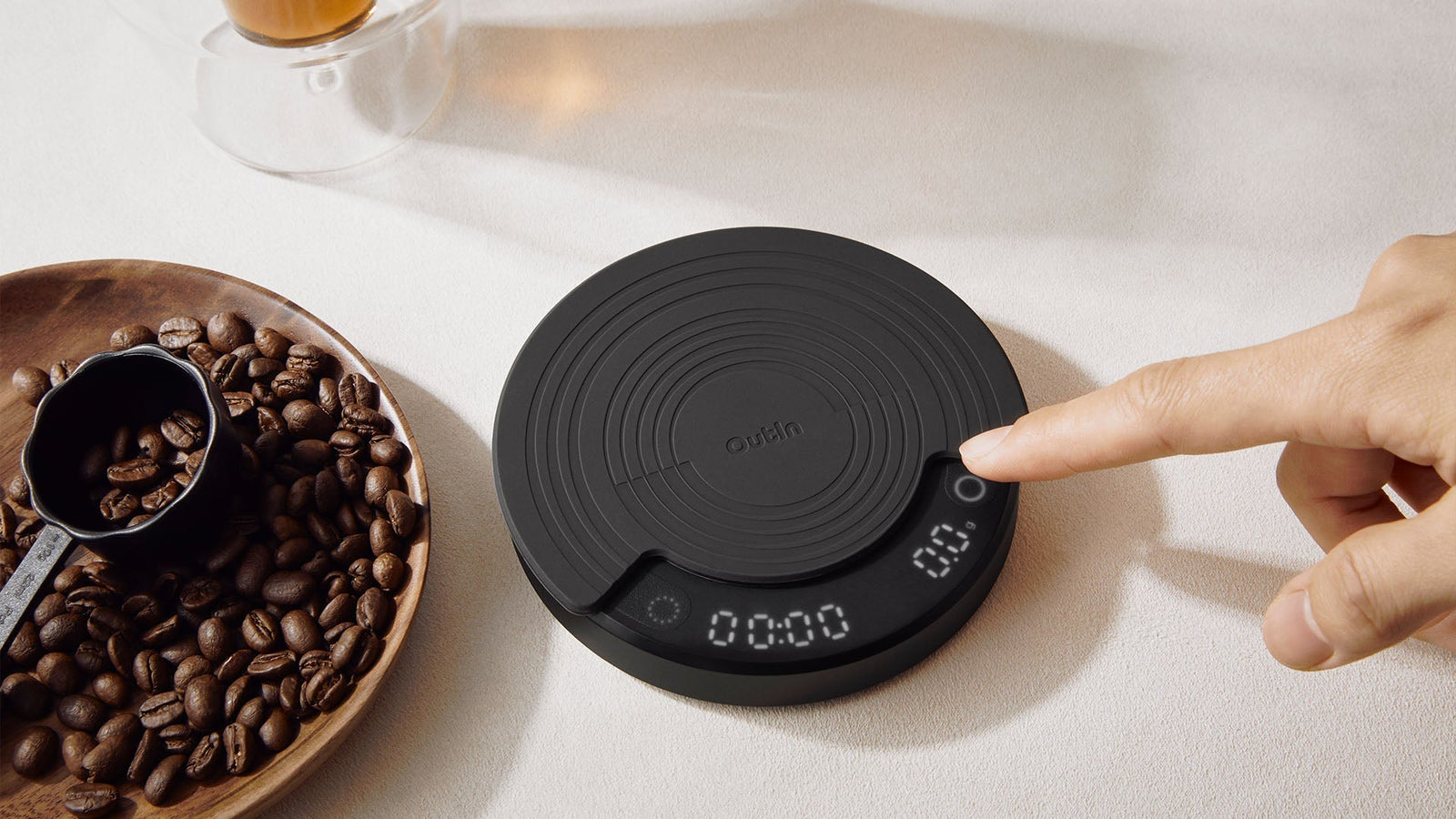 Outln digital coffee scale showing precise measurement with finger pressing button. Coffee beans in wooden bowl nearby for accurate brewing ratio. High-precision scale displays weight for consistent coffee flavor.