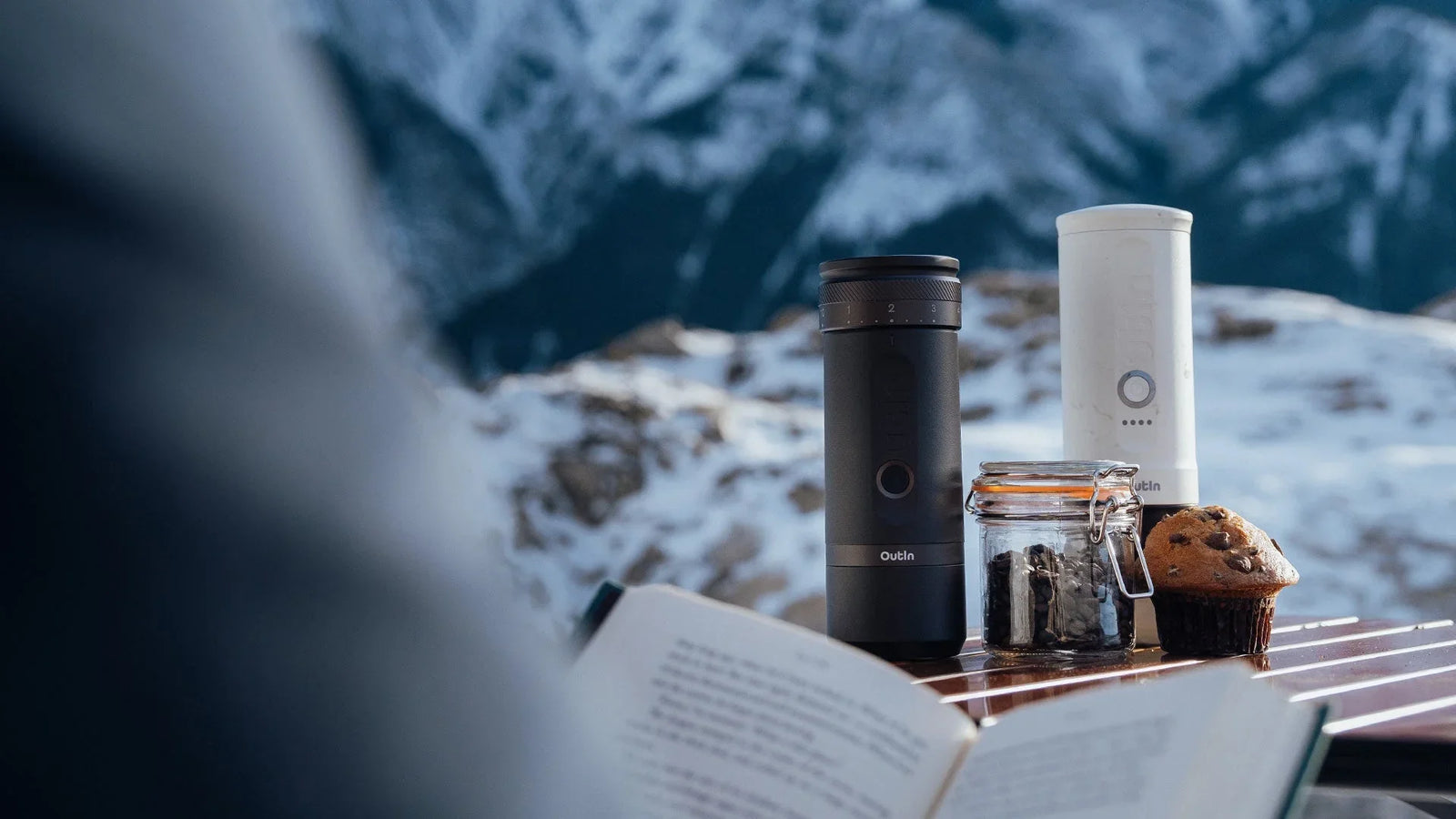 Two portable espresso makers are on a wooden table with an open book in the foreground and a backdrop of snow-covered mountains.