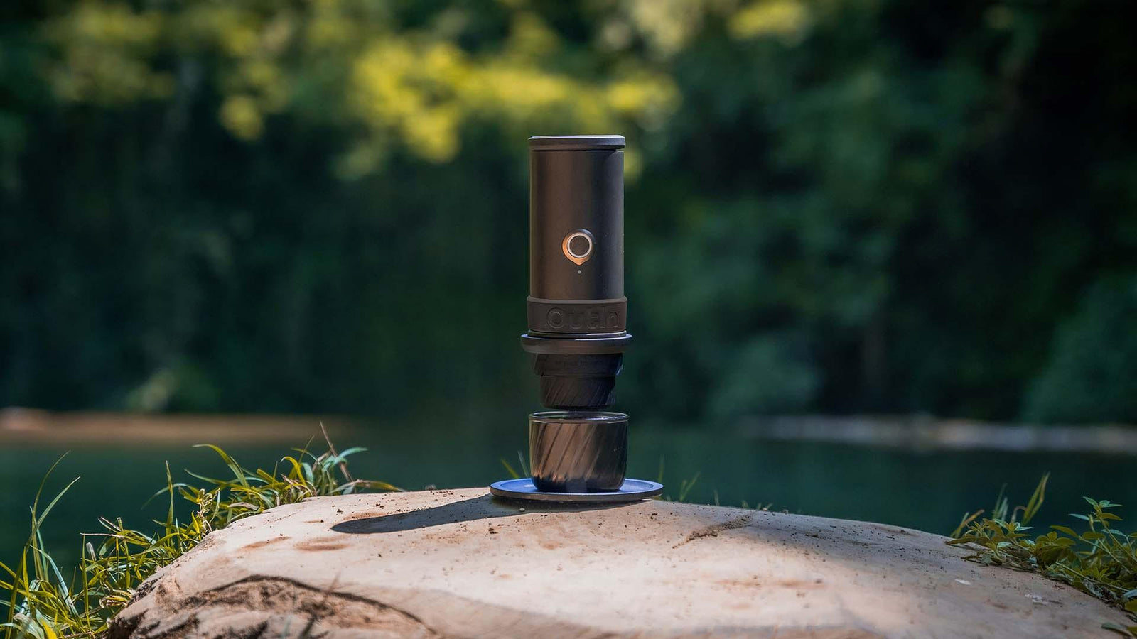 A portable espresso machine brewing a shot into a cup, sitting on a wooden tree stump by a lake.