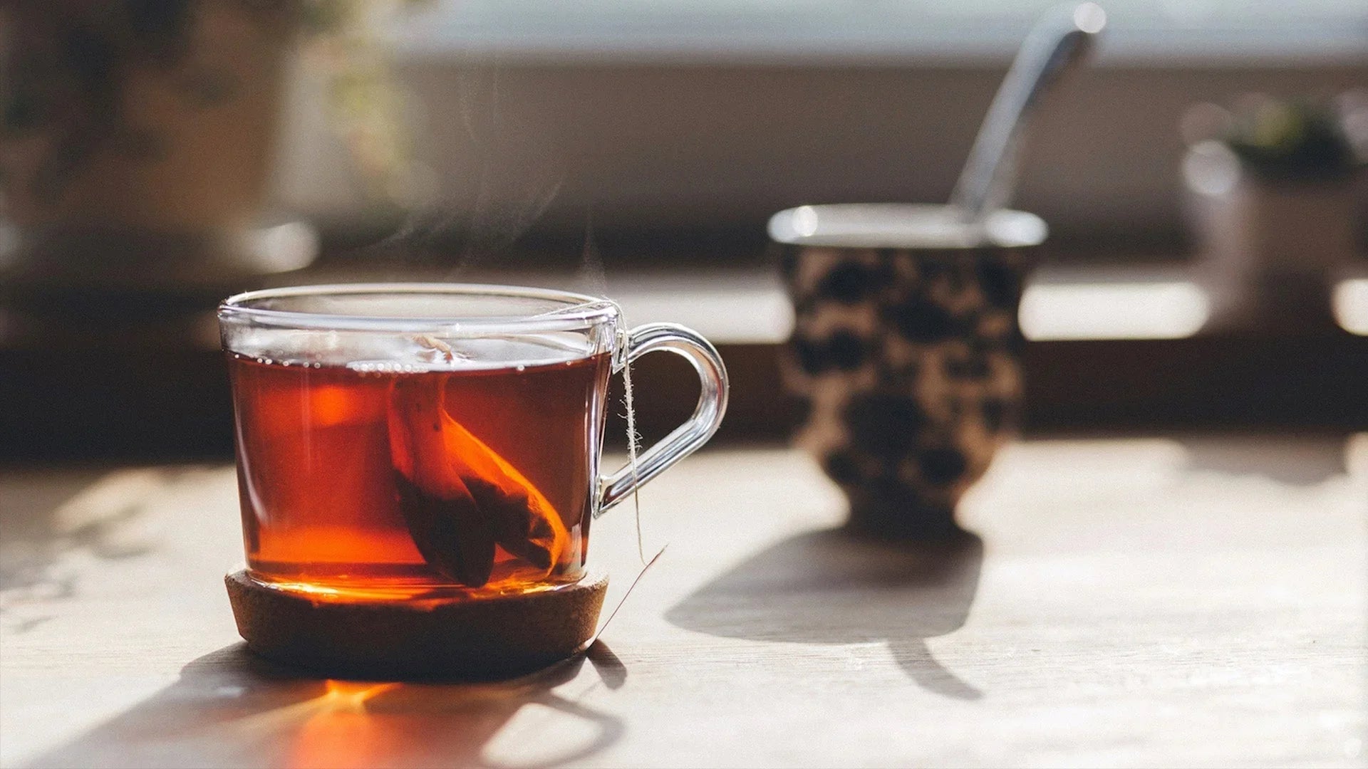 A clear glass cup of steaming hot tea with a tea bag visible, sitting on a cork coaster in warm sunlight, with a blurred second cup in the background creating a cozy atmosphere