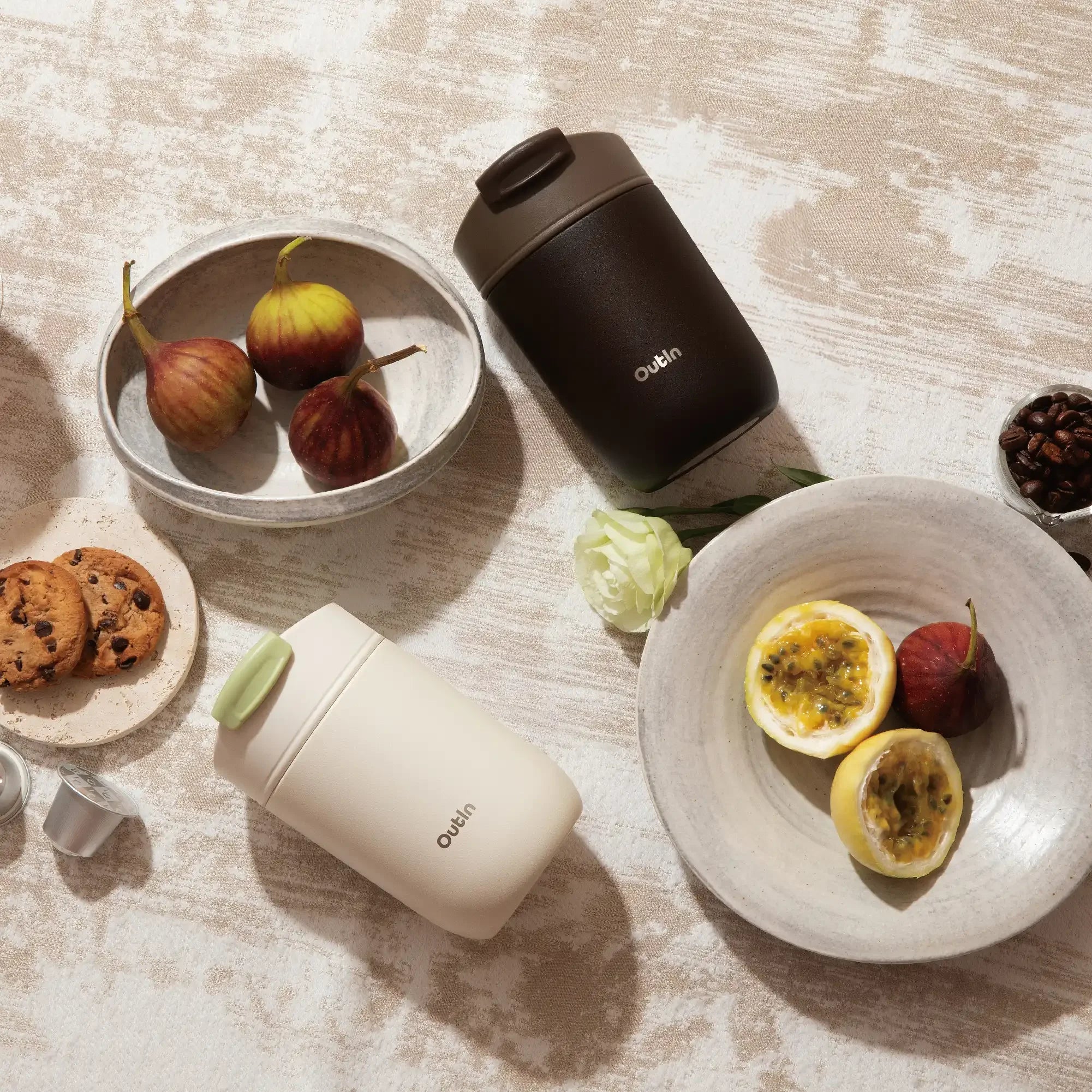 OutIn travel tumblers in dark brown and cream, with figs, passion fruit, cookies, and coffee beans.