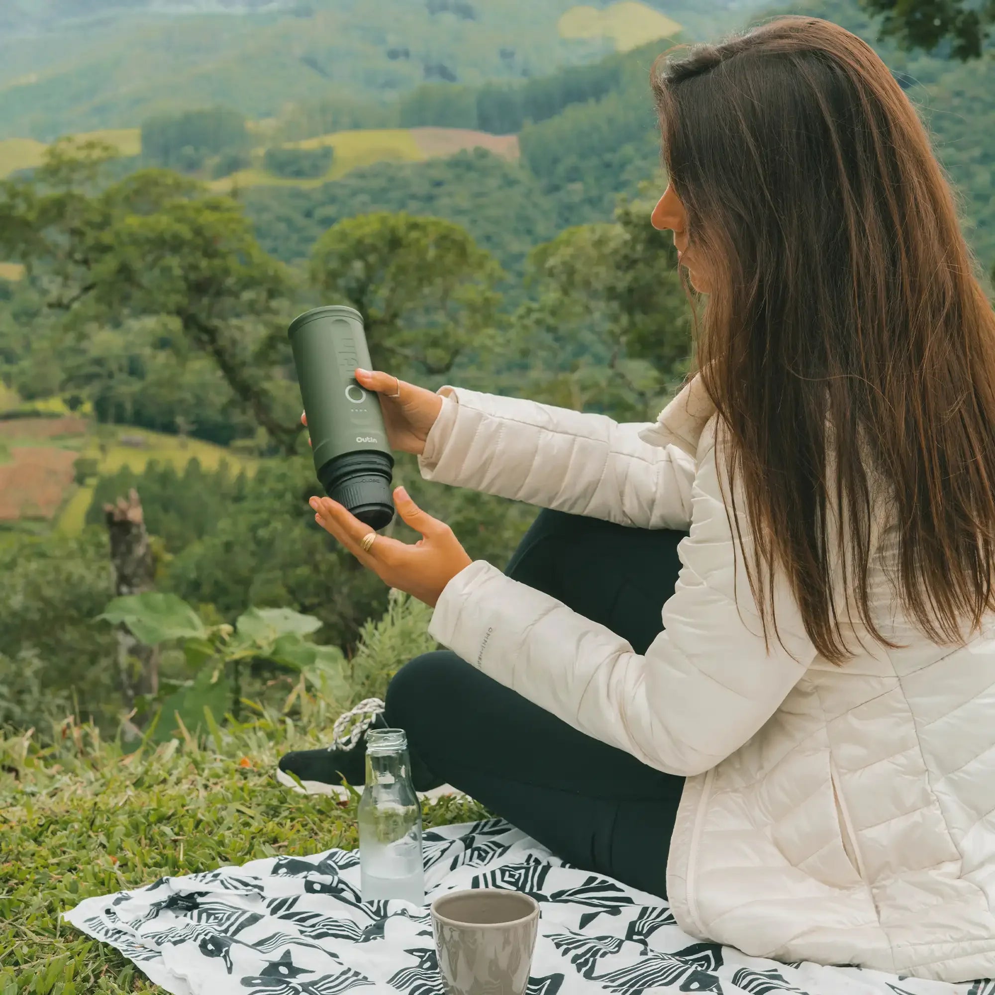 Woman enjoying forest green Nano portable espresso machine outdoors with scenic hilly background.