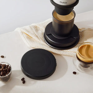 OutIn Claro black coffee scale with textured surface, beside coffee beans, pour-over device, and brewed coffee in a glass.