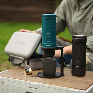 Teal portable espresso machine brewing into mug on black adjustable Universal Coffee Stand outdoors, with grinder and case.