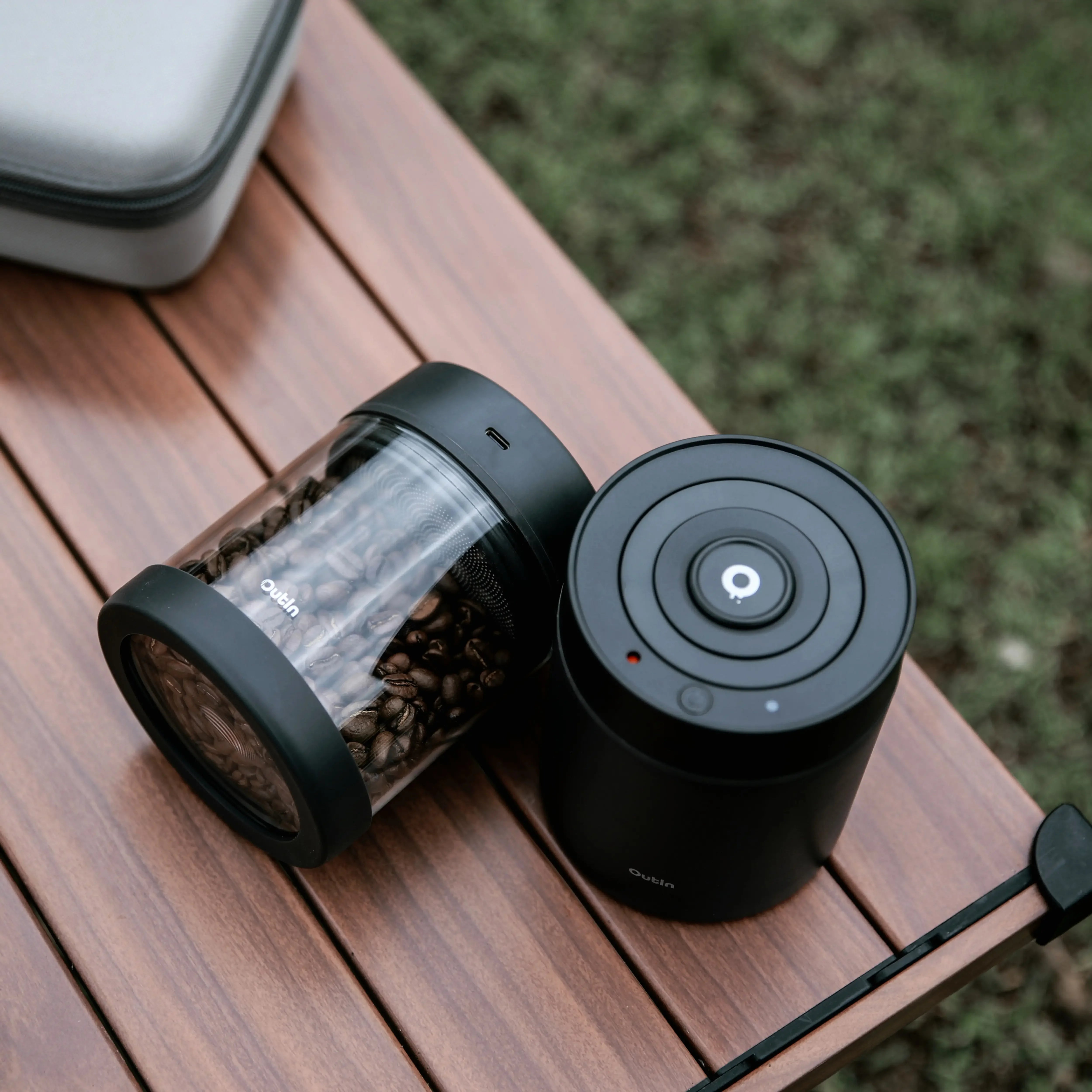 Glass portable vacuum coffee canister filled with beans beside a black electric vacuum coffee canister on a wooden surface.