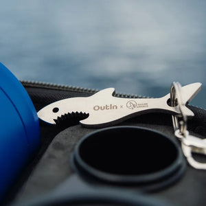 Shark shaped keychain with OutIn x Shark Angels engraving next to ocean blue portable espresso machine.