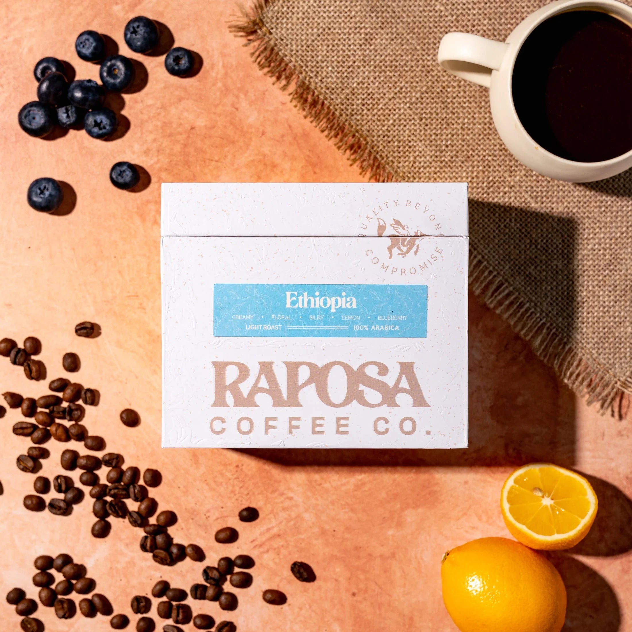Raposa Ethiopia Light Roast coffee beans box with scattered beans, fresh blueberries, lemon, and a coffee cup on burlap.