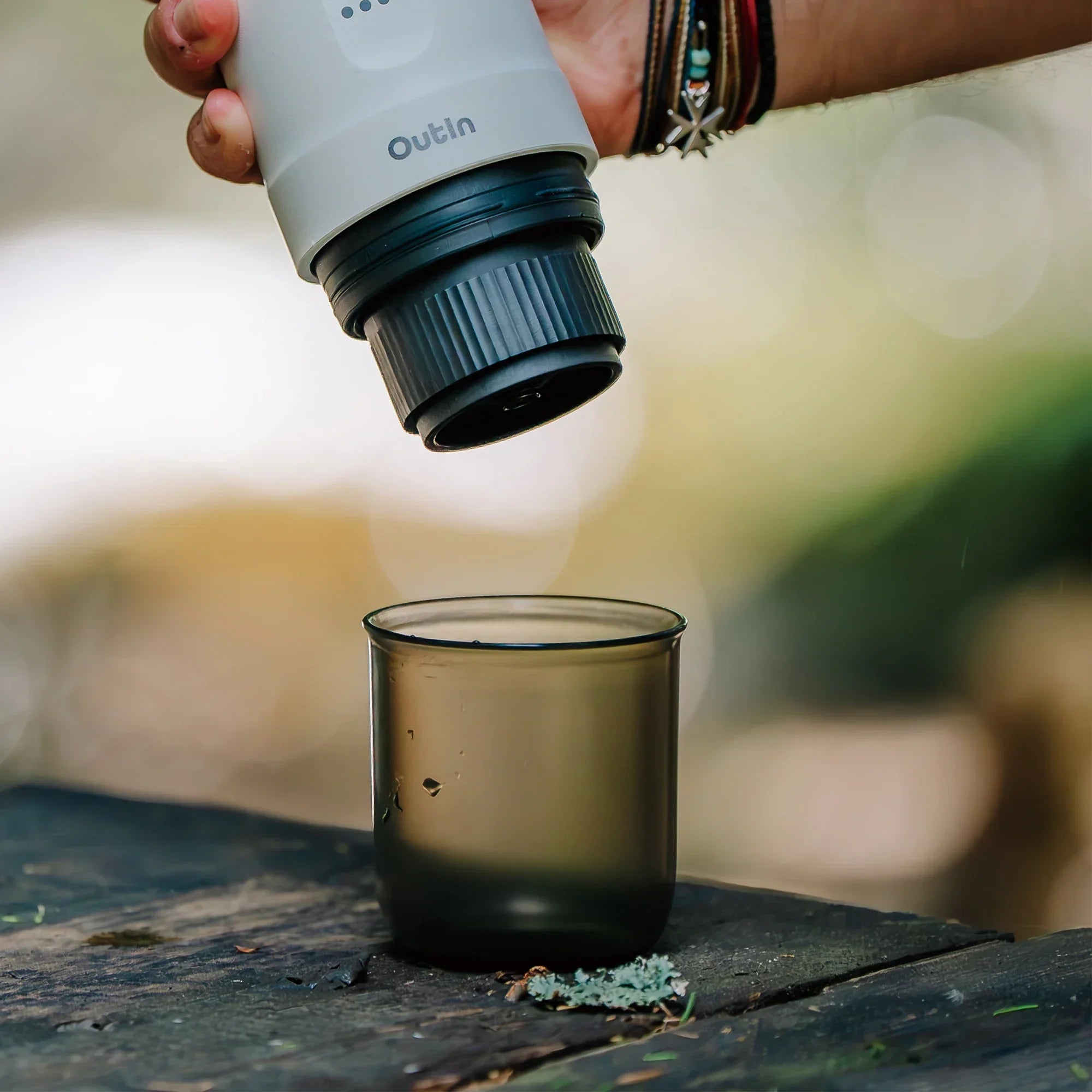 Hand holding OutIn portable espresso machine brewing head over smoky grey BPA-free cup on wooden table for outdoor use.