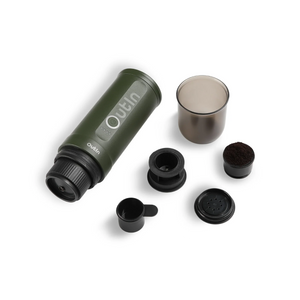Forest green OutIn Nano portable espresso machine with accessories: scoop, grounds basket, capsule adapter, and cup.