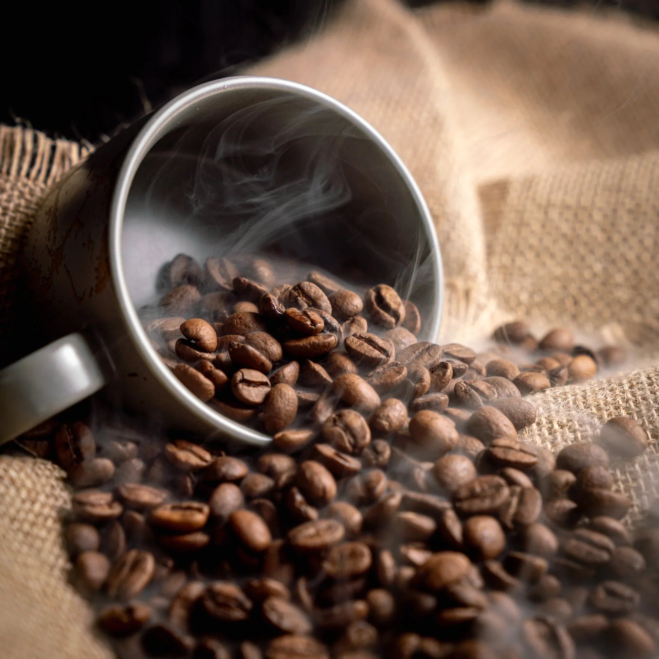 Freshly roasted Ethiopia Yirgacheffe light roast coffee beans spilling from a cup onto burlap, with steam.