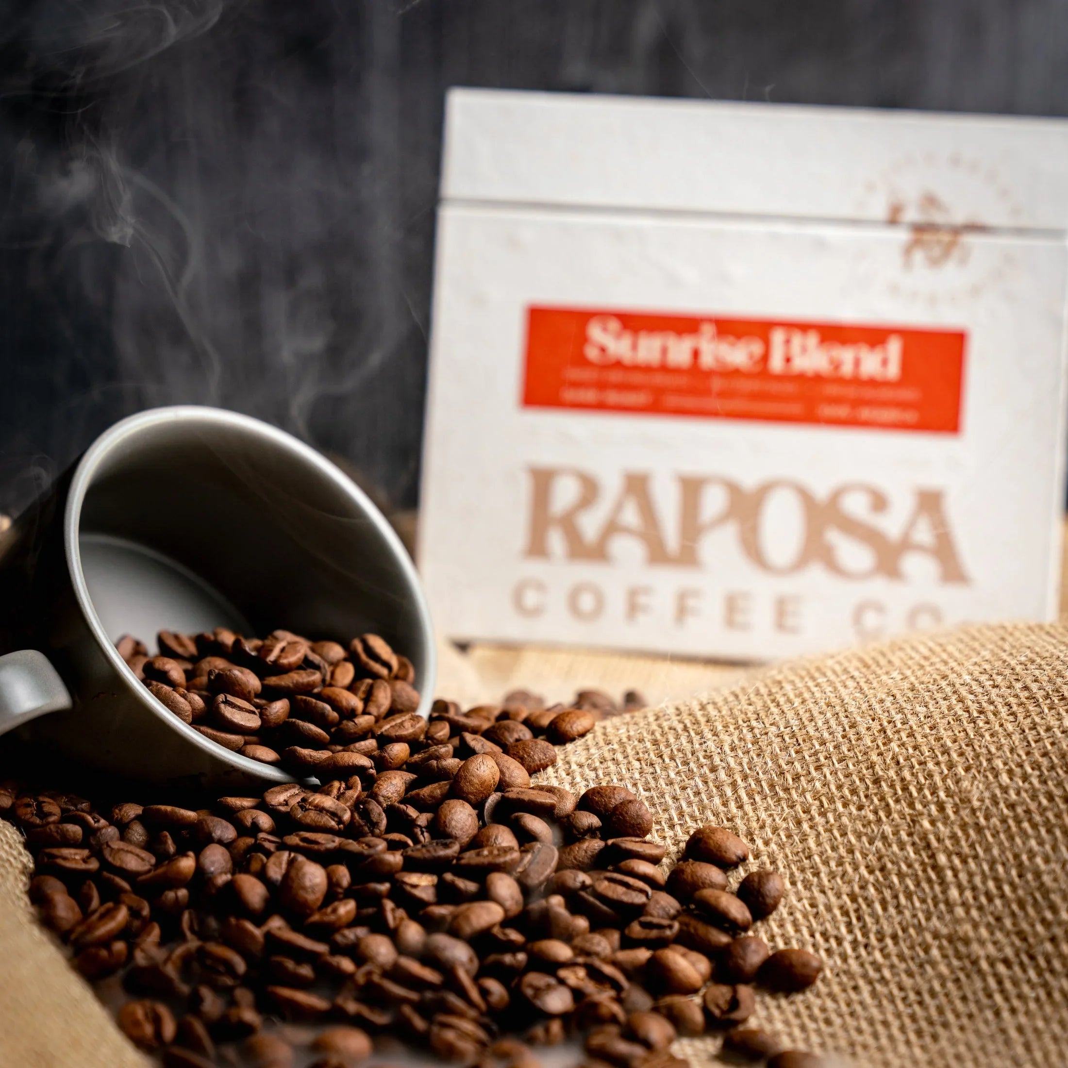 Sunrise Blend dark roast coffee beans spilled from mug onto burlap with Raposa Coffee Co. packaging and steam.