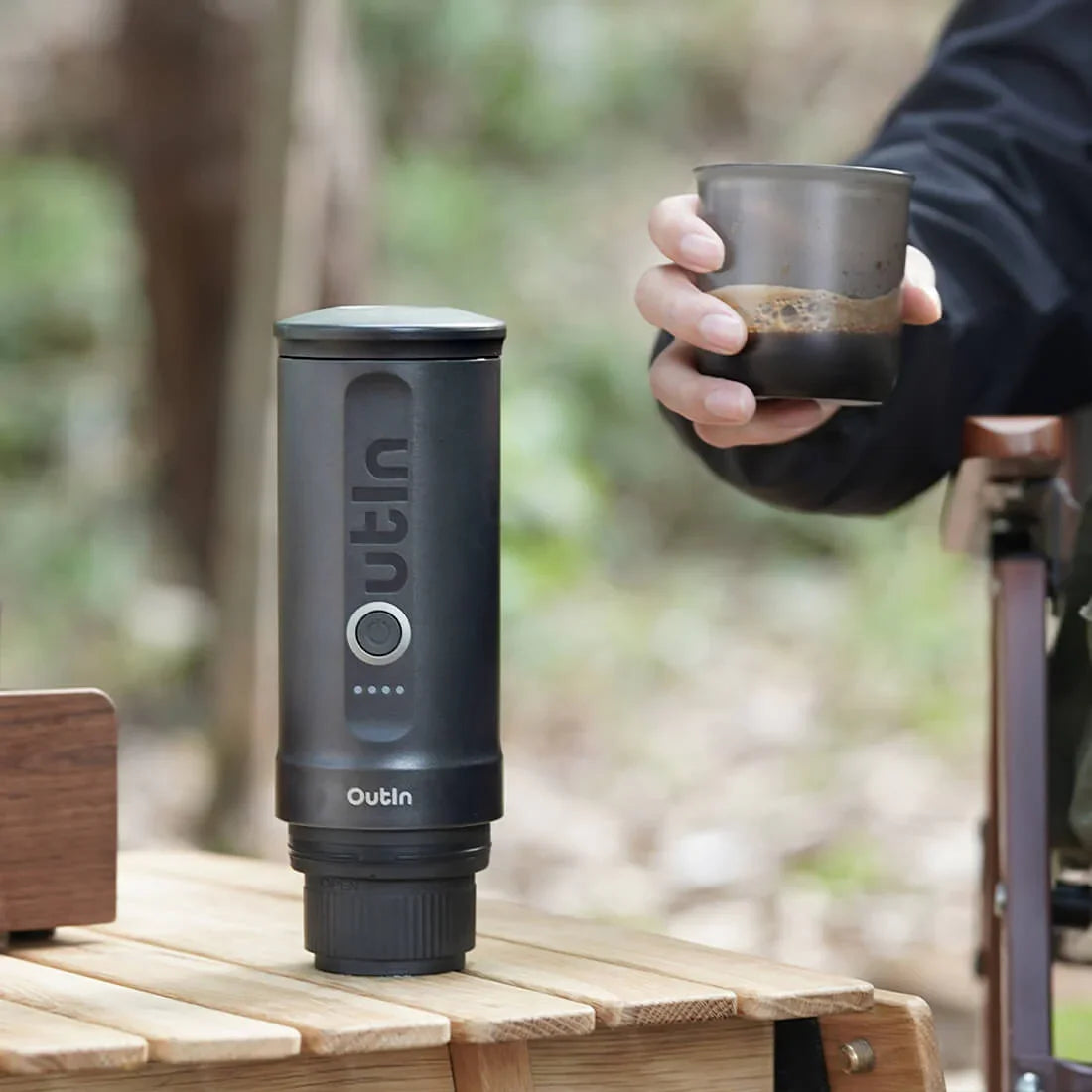 Space grey OutIn Nano portable espresso machine on wooden table, hand holding cup of espresso outdoors.