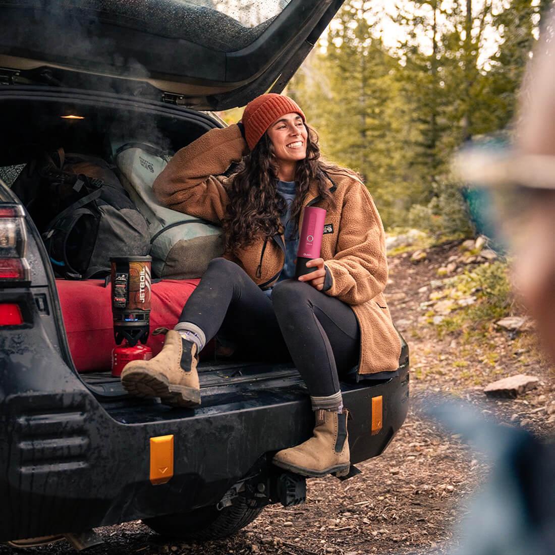Woman with fuchsia Nano portable espresso machine enjoying outdoors near car and camping gear.