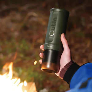Forest green OutIn Nano portable espresso machine held by hand near campfire, showcasing outdoor use.