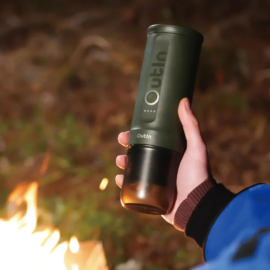 Forest green OutIn Nano portable espresso machine held by hand near campfire, showcasing outdoor use.